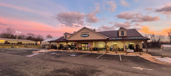 Storefront display featuring a variety of products such as wheelbarrows, plant hooks, seed spreaders, and soil. The foreground shows a lined parking lot, while the background features a beautiful sunset with shades of blue, pink, orange, and yellow.