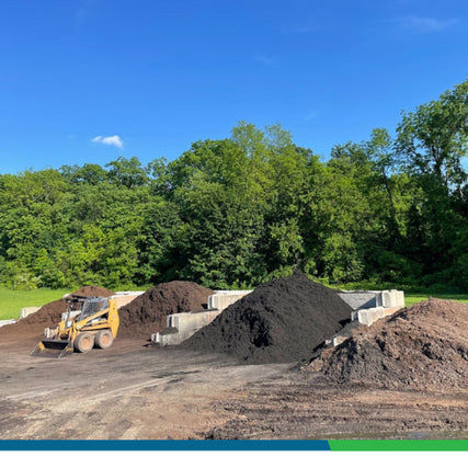 Bagged Mulch & Bulk MulchA yellow skid steer parked in front of a selection of mulch, with shades ranging from light brown to chocolate brown to black. In the background, a variety of green leafy trees can be seen.